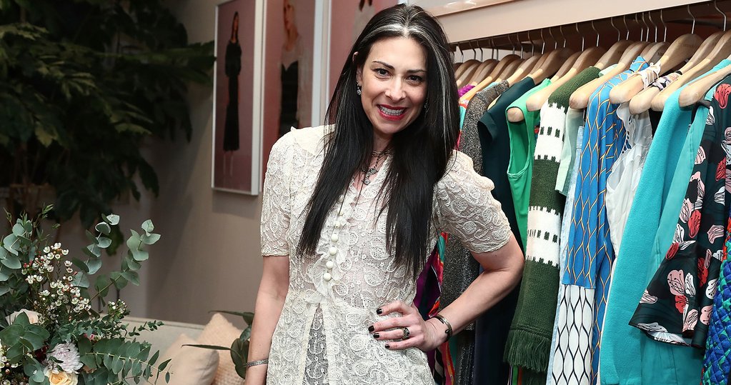Portrait of Stacy London smiling at the camera standing in front of a rolling rack of clothes with one hand on her hip.
